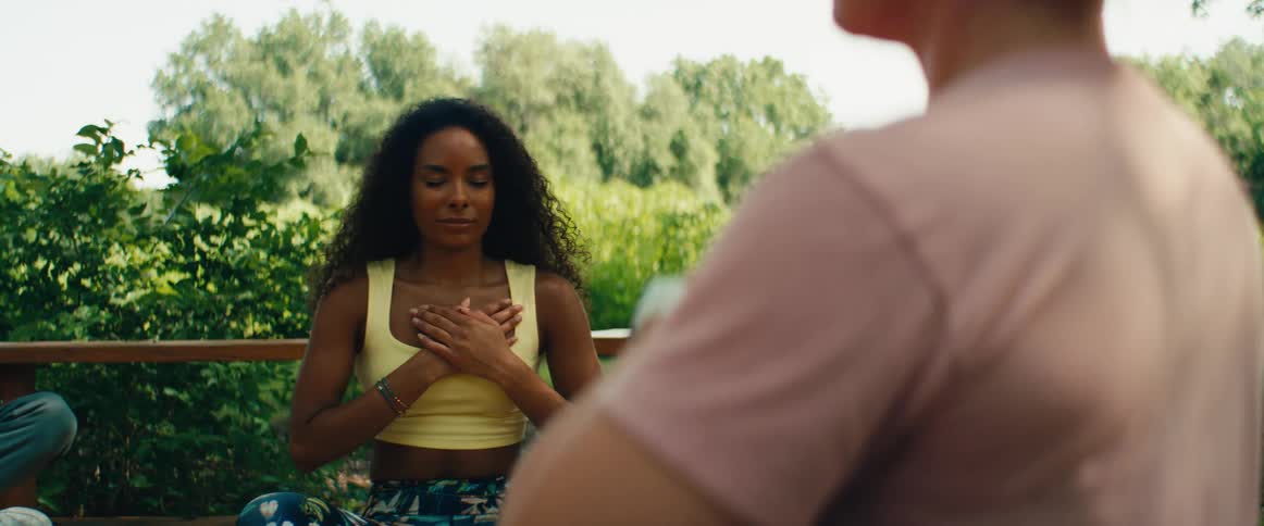 A tranquil woman with eyes closed meditates peacefully outdoors with her hands on her chest, as seen from over her partner's shoulder