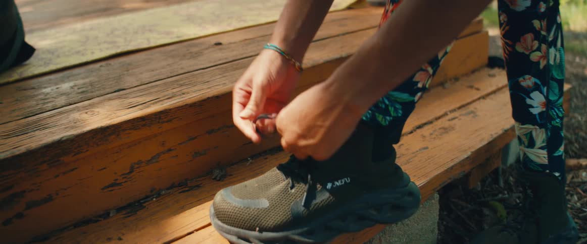 A person wearing colorful floral leggings carefully ties the laces on their green hiking boots while resting their foot on a wooden step outdoors