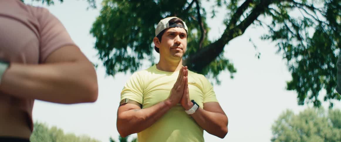 A focused man in a yellow shirt meditates with his hands in a prayer pose while doing yoga outdoors in a park