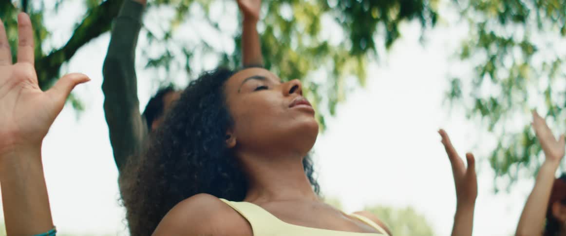 A mindful woman with curly hair participates in an outdoor yoga class, stretching towards the sky in slow motion