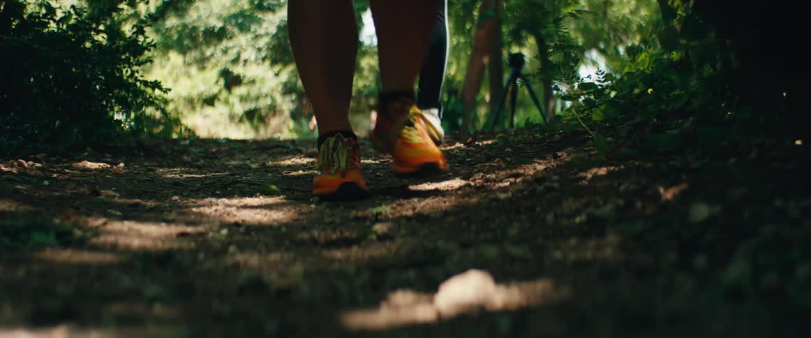 A low-angle slow-motion view shows the feet of a diverse group of people as they walk along a dirt path in a sunlit forest