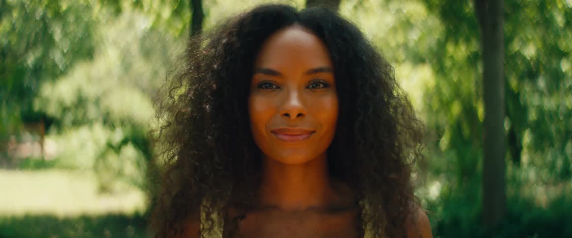 A beautiful woman with curly hair smiles warmly at the camera in a lush green forest