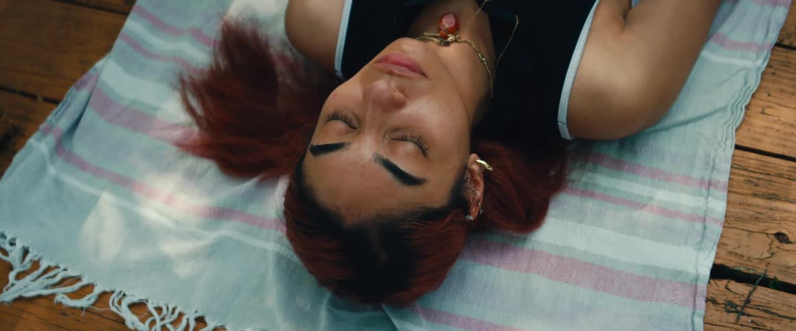 A serene young woman with red hair rests on a blanket outdoors, with dappled sunlight moving across her face