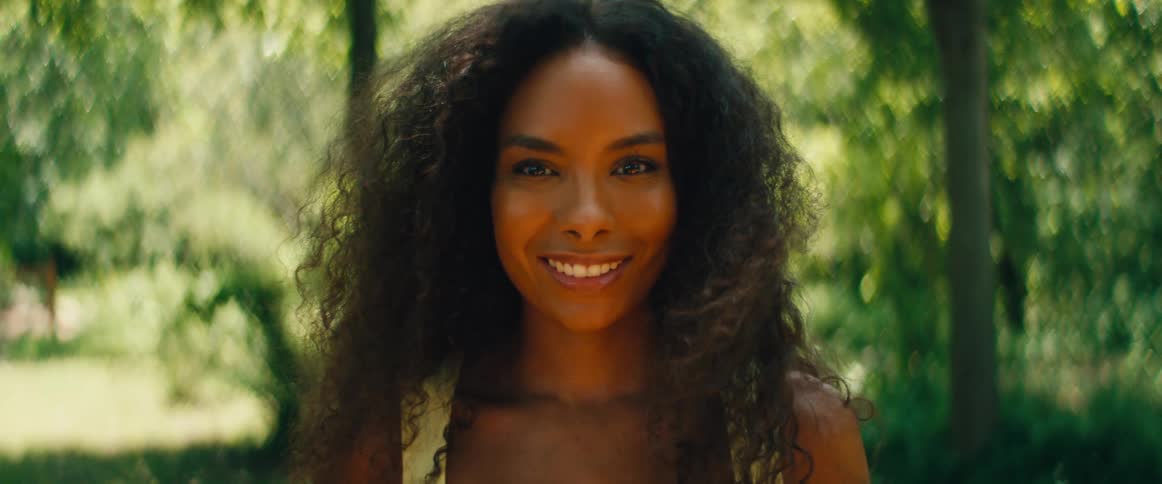 Close-up portrait of a beautiful woman with voluminous curly hair smiling warmly at the camera in a sun-dappled forest