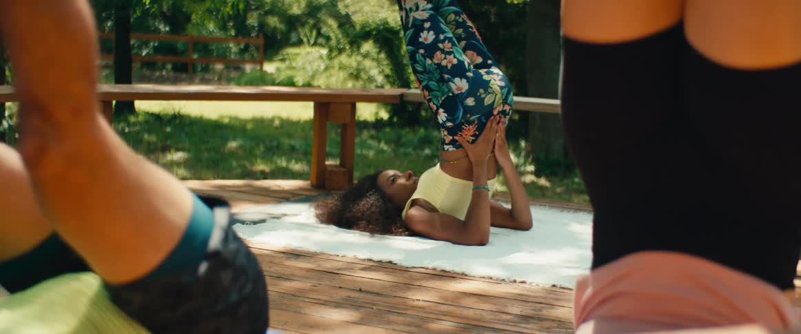 A diverse group of friends holds a supported shoulder stand yoga pose on a wooden deck in a sunlit park