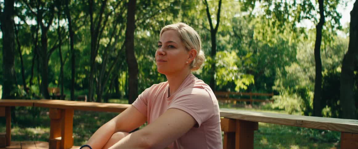 A happy blonde woman with her hair in a bun sits on a wooden bench outdoors and enjoys a pleasant conversation on a sunny day