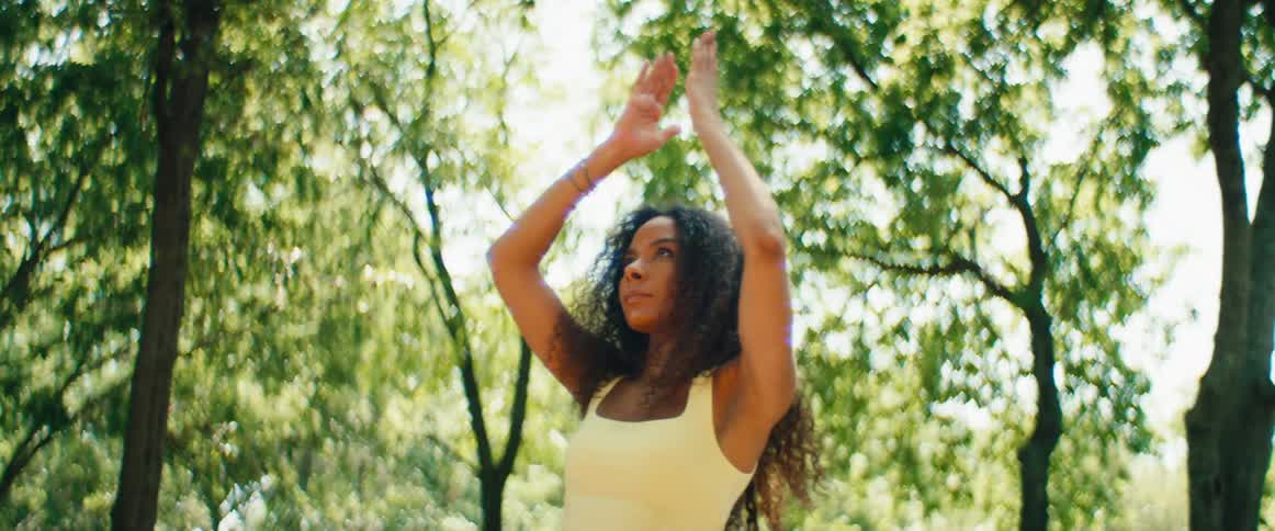 A serene young woman with curly hair performs a slow-motion yoga pose in a sun-dappled park
