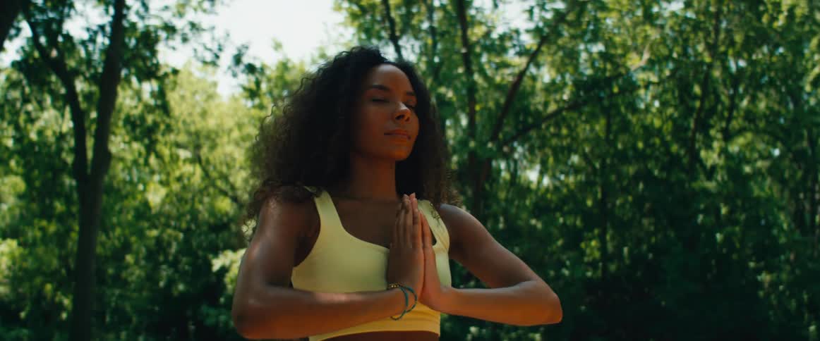 A serene woman with curly hair meditates with her hands in a prayer pose, finding peace in a sunlit park
