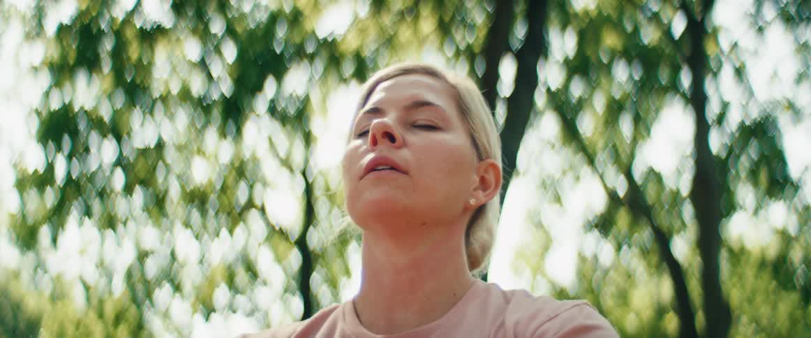A peaceful blonde woman practices mindfulness and deep breathing meditation with her eyes closed in a sun-dappled forest