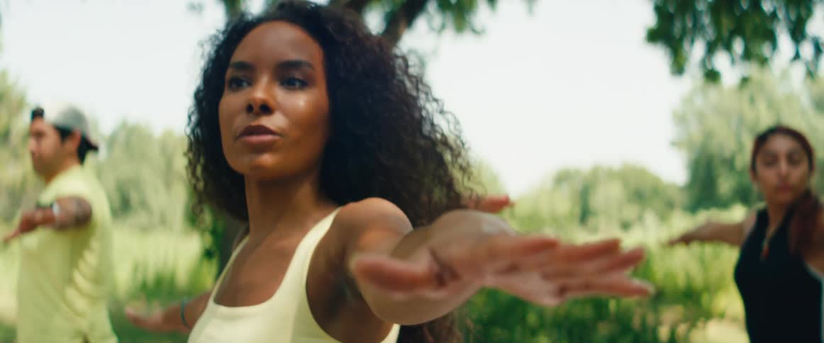 A focused woman with curly hair holds a warrior pose during an outdoor yoga class in a sunny, green park