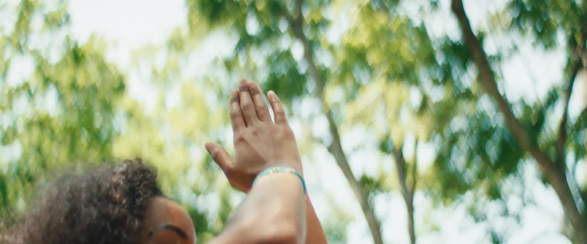 A tranquil woman with curly hair meditates in a sun-dappled park, slowly raising her hands in a prayer pose in an intimate slow-motion close-up