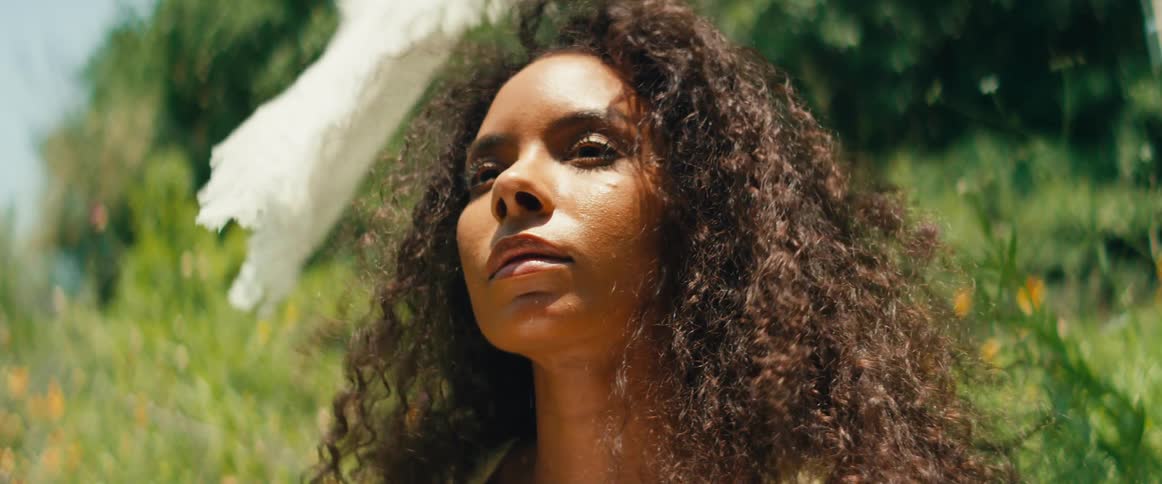 A serene woman with voluminous curly hair sits thoughtfully in a sunlit meadow under a white parasol