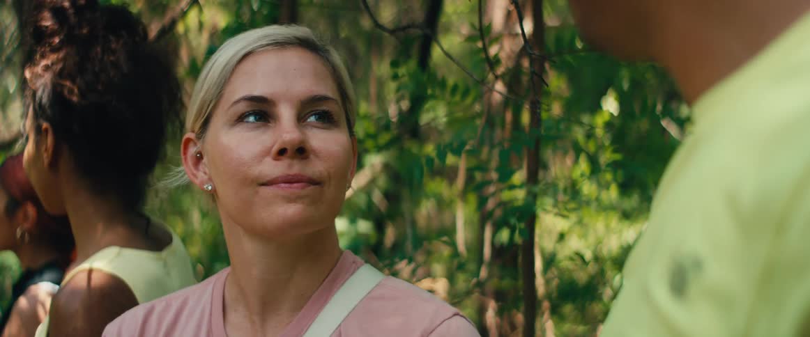 A smiling blonde woman with her hair in a bun listens attentively during a conversation in a sunny forest