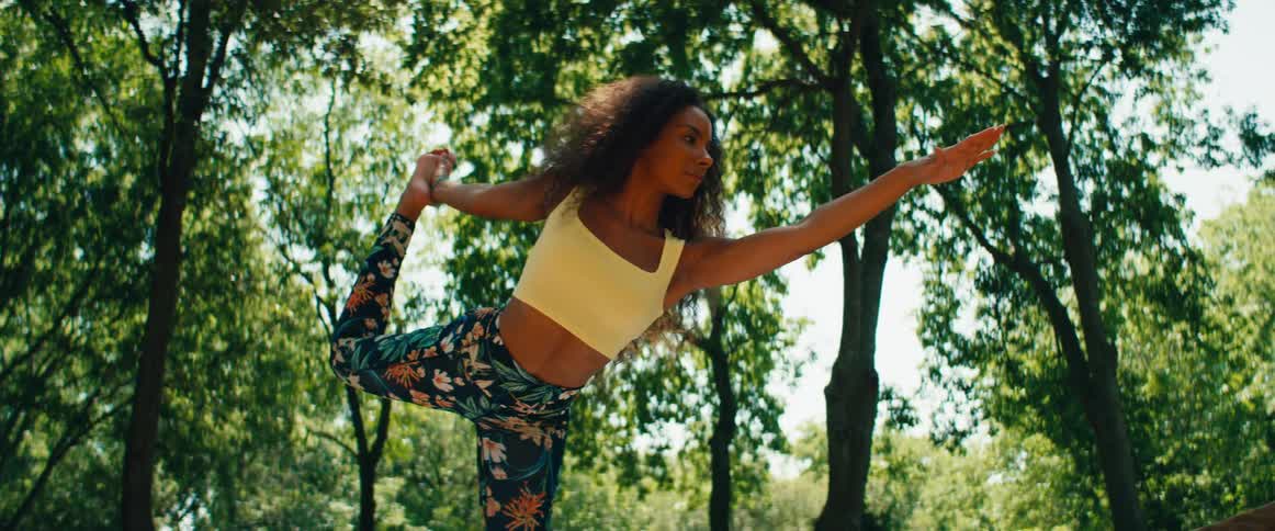 A serene woman performs a balancing yoga pose in a sun-dappled park, captured in slow motion