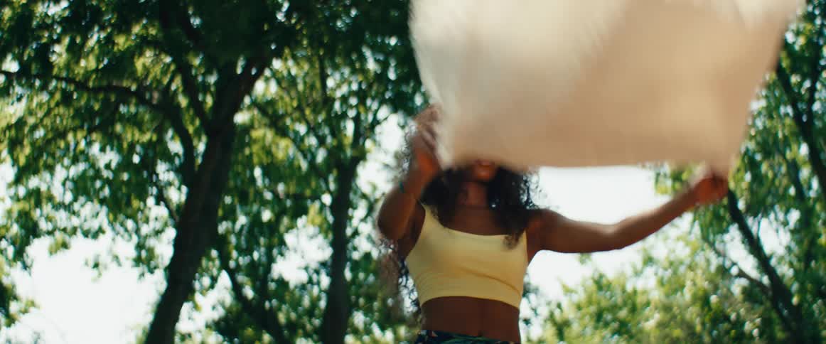A graceful woman with curly hair performs a slow, flowing dance with a white blanket in a sunlit park