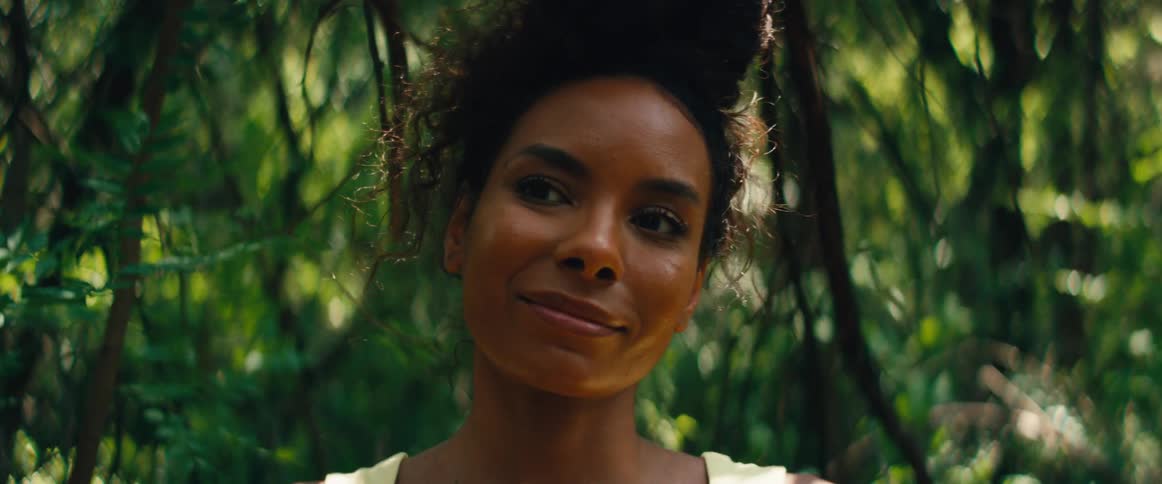 A beautiful woman with curly hair smiles serenely while standing in a lush, sun-dappled forest