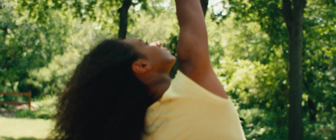 A woman with flowing curly hair gracefully moves through yoga poses in slow motion on an outdoor deck surrounded by lush greenery