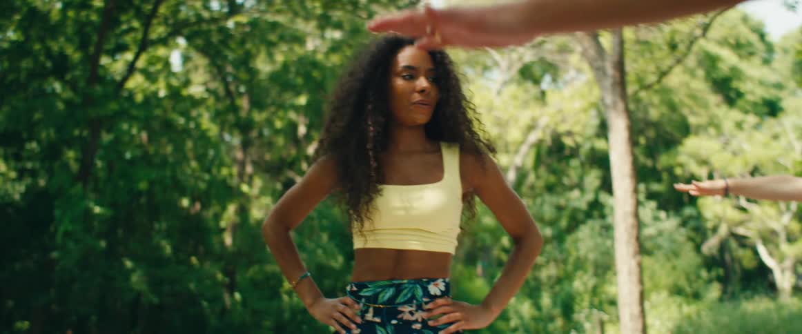 A graceful woman with long curly hair smiles as she leads an outdoor yoga class in a sunlit park in slow motion