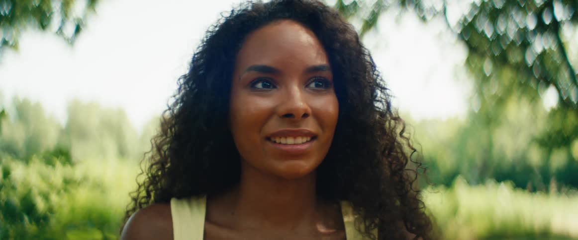 A slow-motion close-up portrait of a beautiful young woman with curly hair smiling on a sunny day in a park