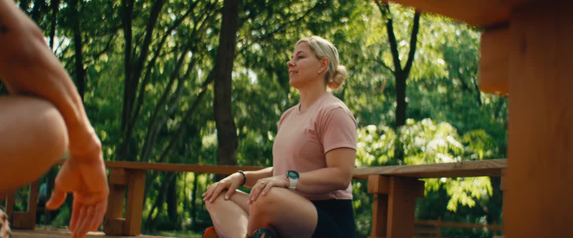 A blonde woman in athletic wear sits cross-legged on a wooden deck and calmly explains something in a serene, green outdoor setting
