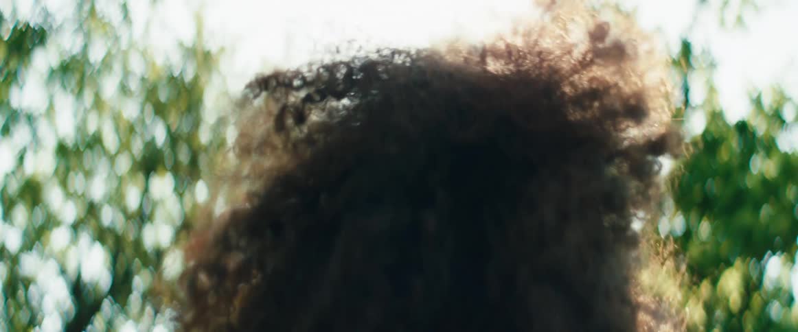 An out-of-focus, slow-motion shot from below of a woman dancing freely with her arms in the air as the sun flares through the trees