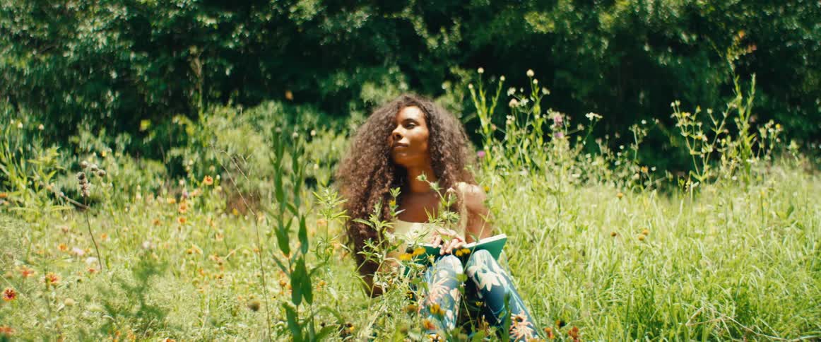 A peaceful woman with curly hair sketches in her notebook while sitting in a vibrant, sunlit meadow of wildflowers