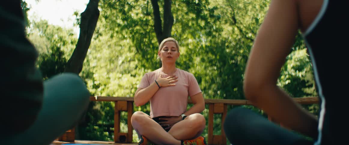 A woman leads a meditation and breathing exercise while sitting in a lotus position on a wooden deck surrounded by tranquil nature