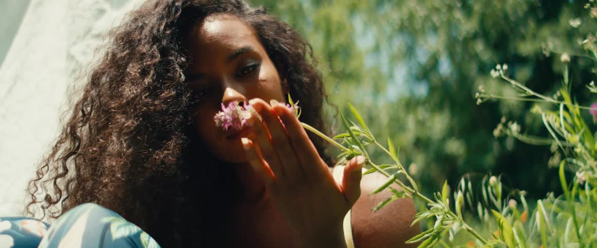 A serene Black woman with curly hair gently smells a purple wildflower in a lush, sun-drenched garden in slow motion