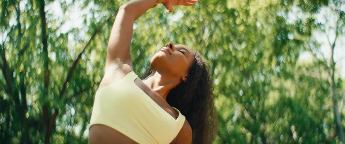A serene woman with beautiful curly hair gracefully practices yoga stretches outdoors in slow motion, surrounded by lush green nature