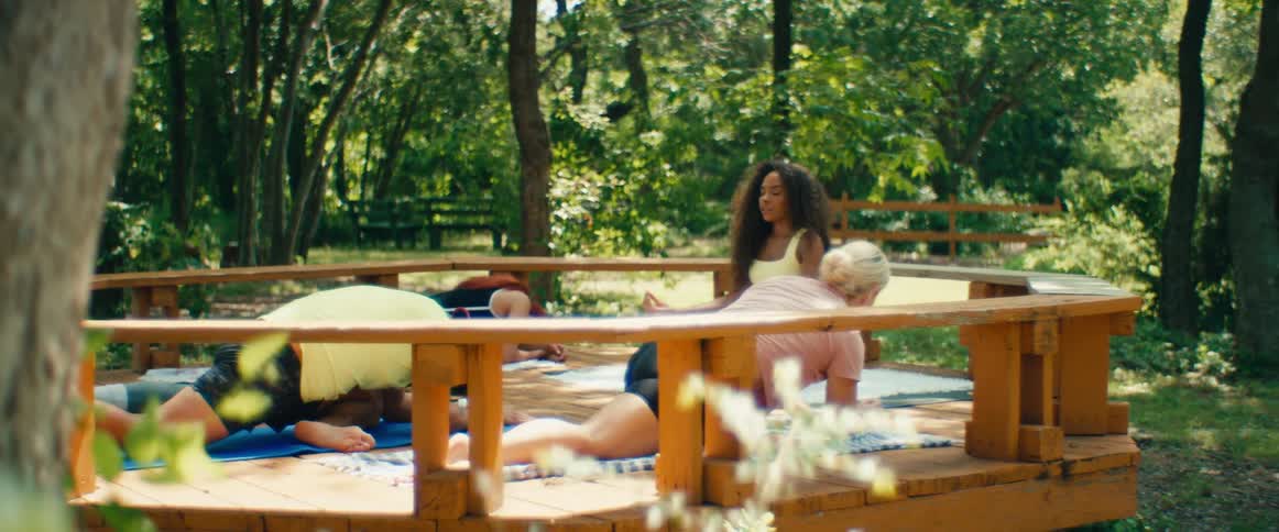 A yoga instructor leads a diverse class through poses on a wooden deck in a serene, sunny park