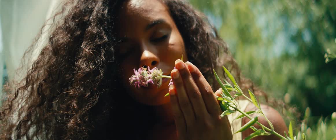 A serene woman with curly hair gently smells a purple wildflower in a sunlit garden in slow motion