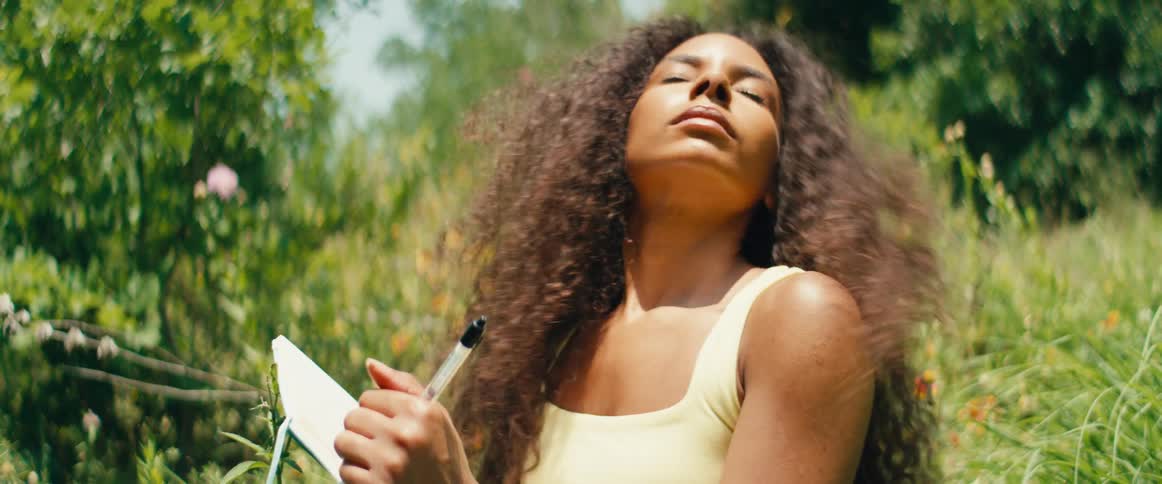 A beautiful young woman with curly hair journals in a sun-drenched garden, pausing to lift her face to the warm sunlight