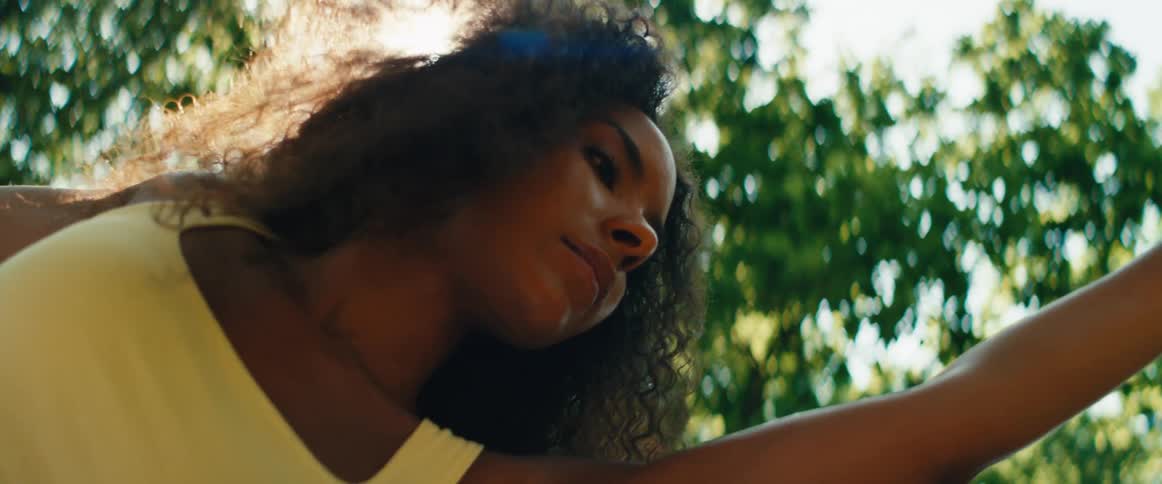 A radiant Black woman with flowing curly hair basks in the warm sunlight, moving gracefully amidst lush green trees in slow motion