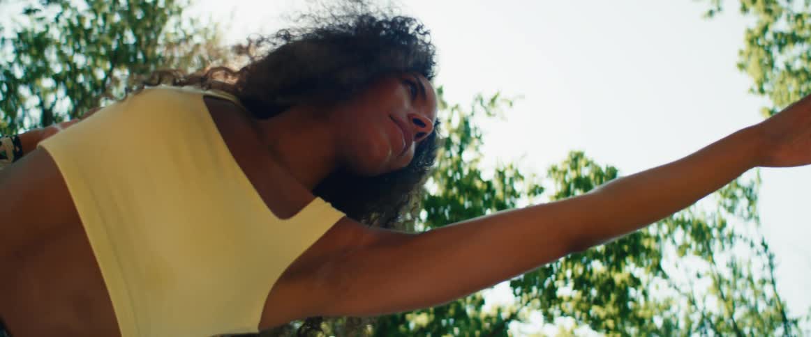A low angle slow motion shot of a woman with curly hair holding a balancing yoga pose in a park as the sun shines brightly behind her