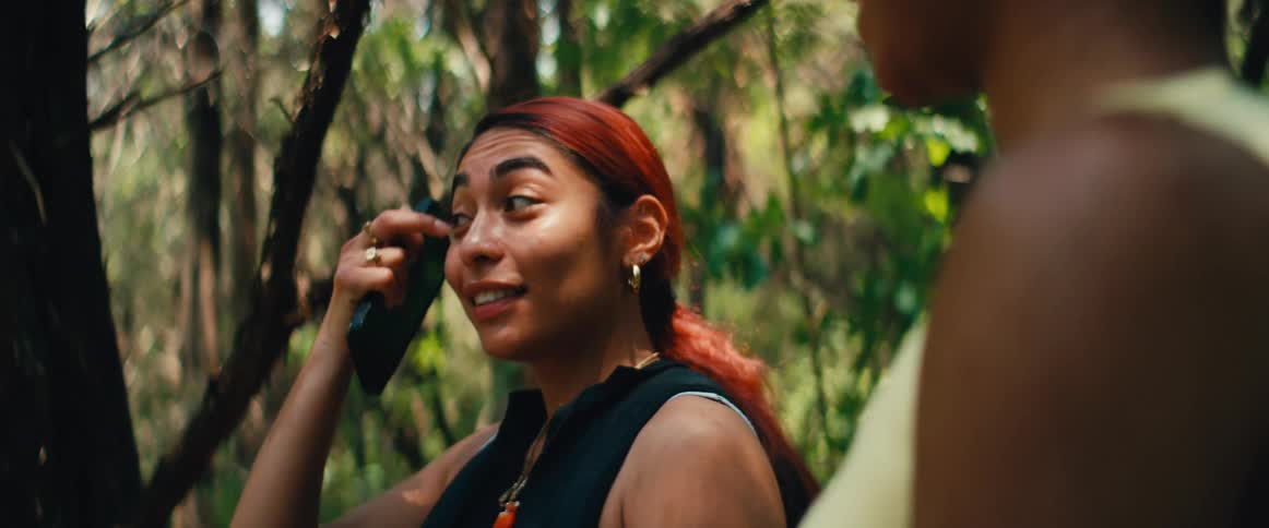 A young woman with dyed red hair smiles and talks with a friend while hiking through a sunlit forest