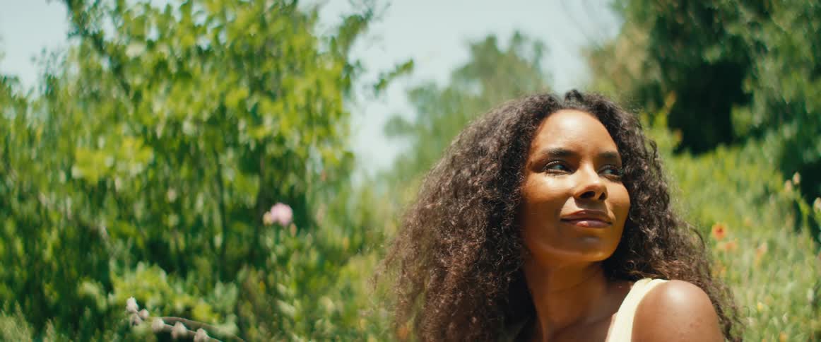 A beautiful woman with curly hair closes her eyes to enjoy the warm sunlight in a peaceful garden setting