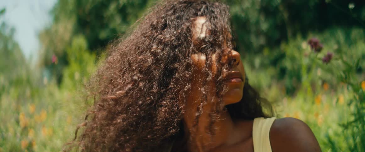 A thoughtful woman with curly hair writes in a journal, relaxing in a sunlit garden as the breeze blows through her hair