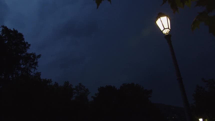Lightning flashes illuminate the dark, cloudy sky behind a glowing street lamp on a stormy night