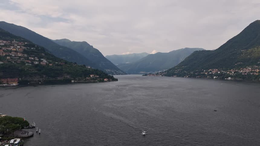 A serene aerial pullback shot reveals the stunning landscape of Italy's Lake Como, surrounded by majestic mountains under a cloudy sky