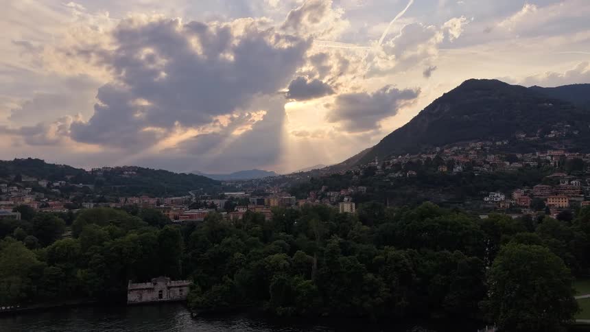Aerial flight over a tranquil lake towards a scenic European city with sunbeams illuminating the sky at dusk