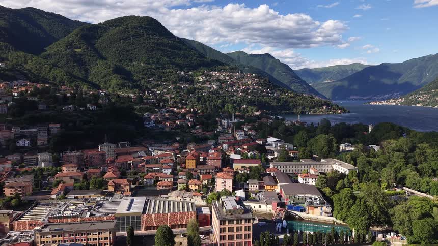 A slow aerial push-in shot captures the stunning landscape of a town nestled between green mountains and a beautiful lake in Italy