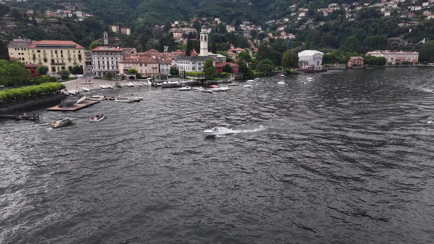 A white motorboat cruises across the lake in front of the picturesque town of Cernobbio, nestled at the base of the mountains in Lake Como, Italy