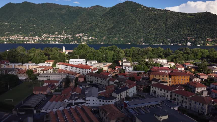 An aerial view slowly advances over the picturesque Italian town of Como, with the stunning Lake Como and green mountains in the distance