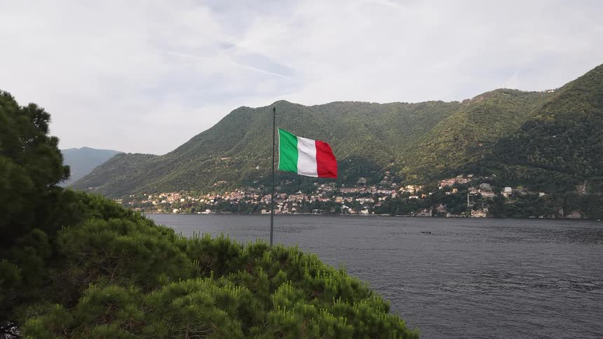 An aerial view slowly approaches the iconic Italian flag waving in the breeze over Lake Como with a mountain village in the background