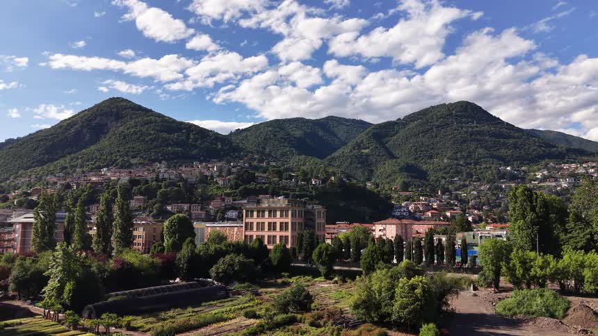 An aerial pullback shot reveals a town nestled at the base of lush green mountains under a partly cloudy sky