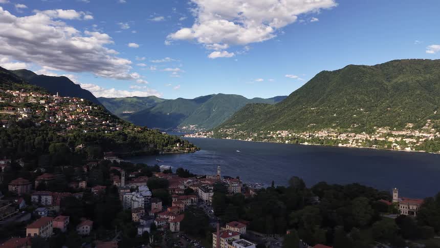 Slow backward aerial shot over a charming Italian town on the shores of Lake Como, surrounded by majestic green mountains under a blue sky