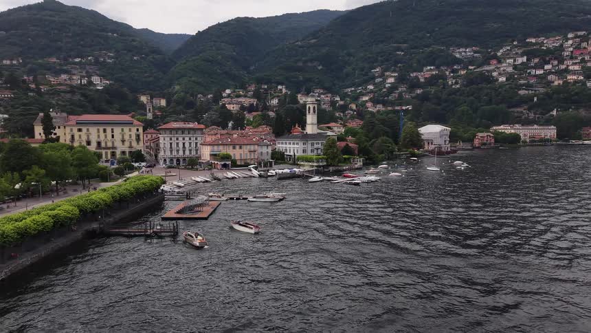 A serene, wide view of the lakeside town of Cernobbio, Italy, with boats moored in the harbor and villas climbing the lush green hillsides of Lake Como