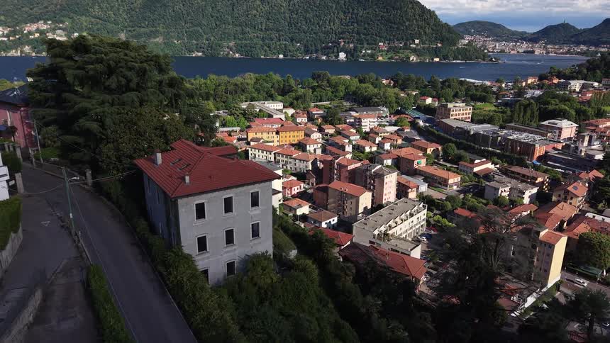 A slow aerial push-in shot reveals a charming Italian town nestled on the picturesque shores of a lake, surrounded by lush green mountains