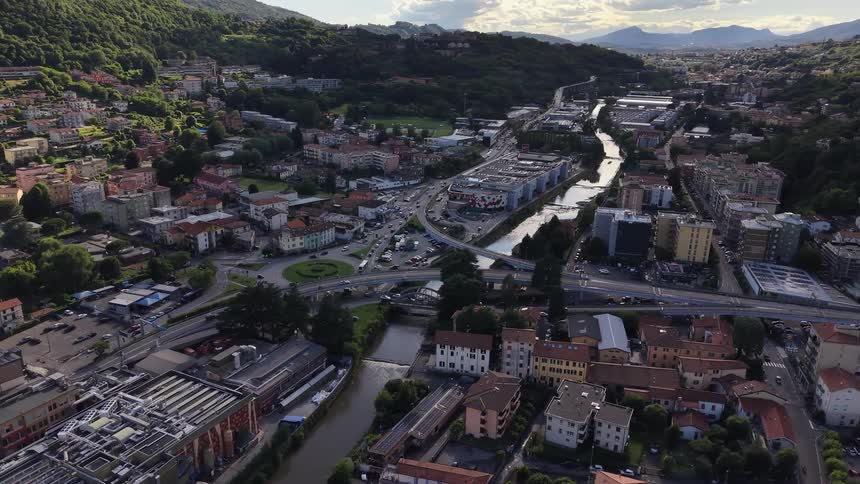 Aerial pull-back shot reveals a picturesque European town nestled between green hills, with a river and a busy road interchange