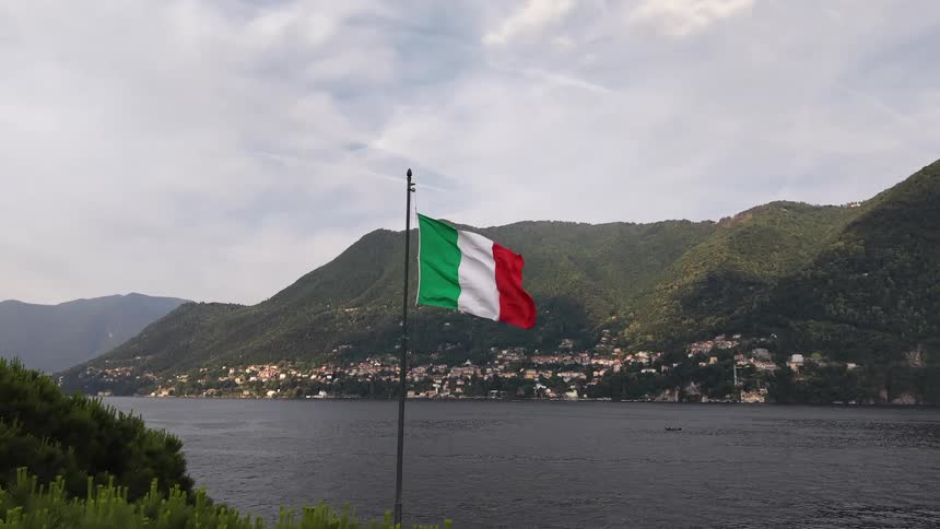 An aerial view pulls back from a waving Italian flag to reveal the stunning mountain and lake landscape of Como, Italy